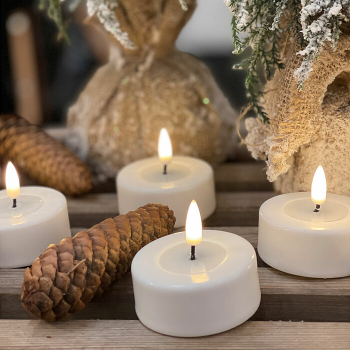 Vier brennende weiße Teelichter von Deluxe Homearts stehen auf einem Holzbrett. Um die Teelichter herum sind dekorativ Tannenzapfen und schneebedeckte Zweige arrangiert. Im Hintergrund befinden sich weitere winterliche Dekorationen.