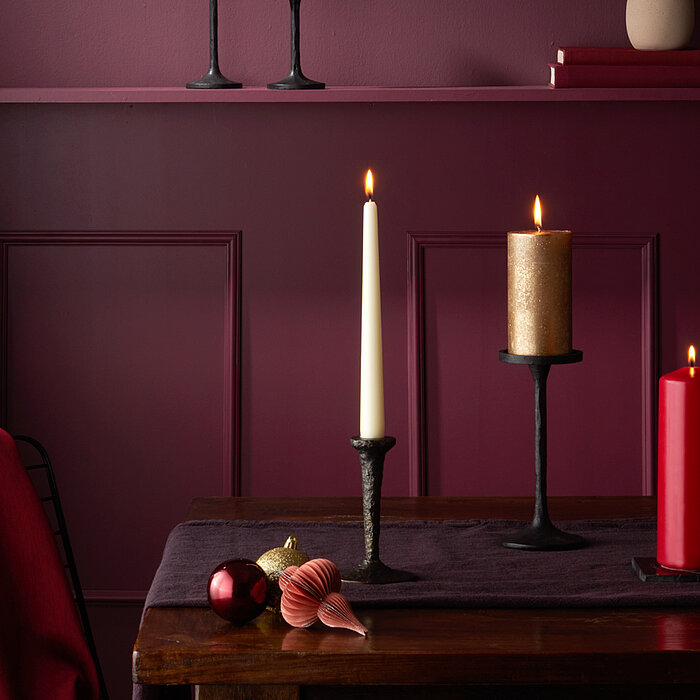 In dem Bild sind vier brennende Bolsius Tafelkerzen zu sehen. Die Kerzen stehen in schwarzen Kerzenhaltern auf einem dunkelvioletten Tuch, das auf einem Holztisch liegt. Zwei Kerzen sind rot, eine ist weiß und eine weitere ist goldfarben. Im Hintergrund s