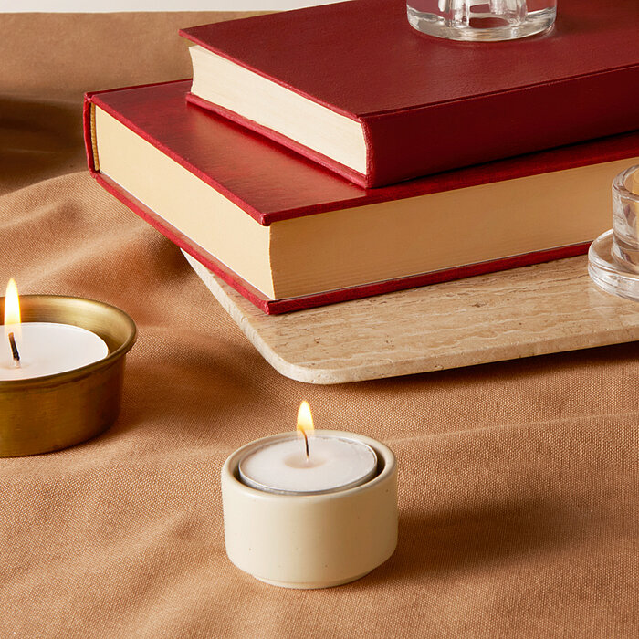 Zwei brennende Bolsius Teelichter in Haltern stehen auf einem beigen Tuch. Eines der Teelichter befindet sich in einem goldenen Halter und das andere in einem weißen Halter. Im Hintergrund liegen zwei rote Bücher auf einem Marmorbrett, daneben steht ein w