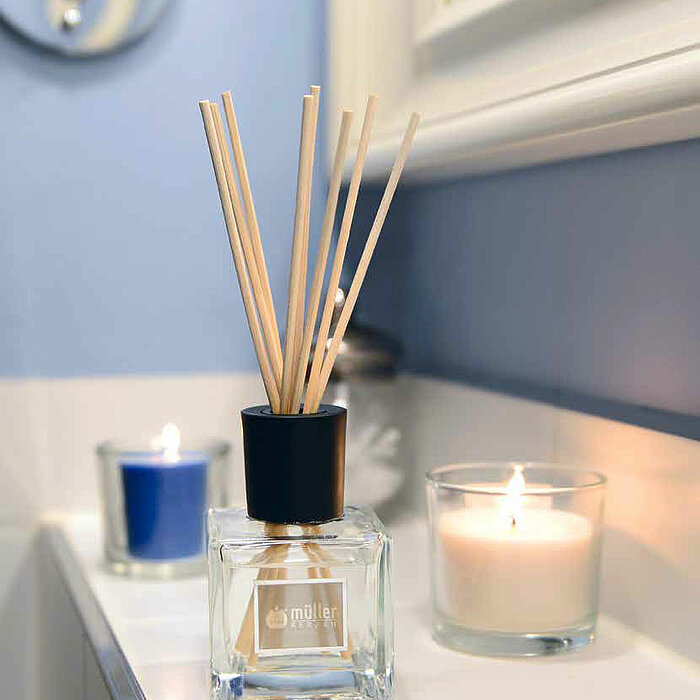 Ein Raumduft von Müller in einem quadratischen Glasbehälter mit mehreren Holzstäbchen steht auf einer weißen Ablage. Daneben sind zwei brennende Kerzen in einfachen Glasbehältern platziert, eine in Blau und eine in Weiß. Der Hintergrund ist in verschieden