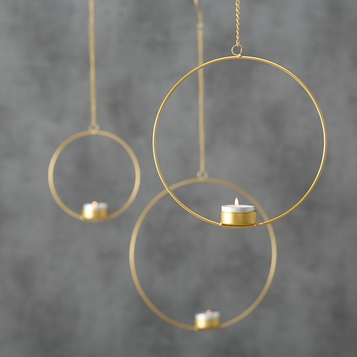 Drei kleine, brennende Teelichter von Boltze schweben in minimalistischen Metallringen, die an Ketten von der Decke hängen. Die Ringe sind in unterschiedlicher Höhe arrangiert, mit einem grauen, unscharfen Hintergrund, der eine ruhige und moderne Atmosphä