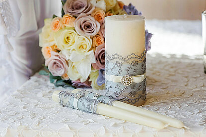 Auf einem zart durchsichtigen, mit bestickten weißen Seiden-Tischtuch liegen zwei elfenbeinfarbene Spitzkerzen, die mit einer Banderole aus einem zarten Spitzentuch verziert sind. Dahinter steht eine elfenbeinfarbene Stumpenkerze, deren Banderole ebenfalls aus einem zarten, spitzenverzierten Tuch besteht. Neben den Kerzen liegt ein wunderschöner, bunter Strauß mit Rosen. Im Hintergrund erkennt man einen Sessel, der mit einem weißen Tüllstofftuch bedeckt ist. Die Szene ist hell und strahlt eine romantische, verträumte Atmosphäre aus.